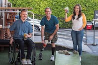 Three friends playing cornhole outdoors, enjoying drinks and smiling facing the camera. One is seated in a wheelchair, another is wearing shorts revealing a prosthetic leg.