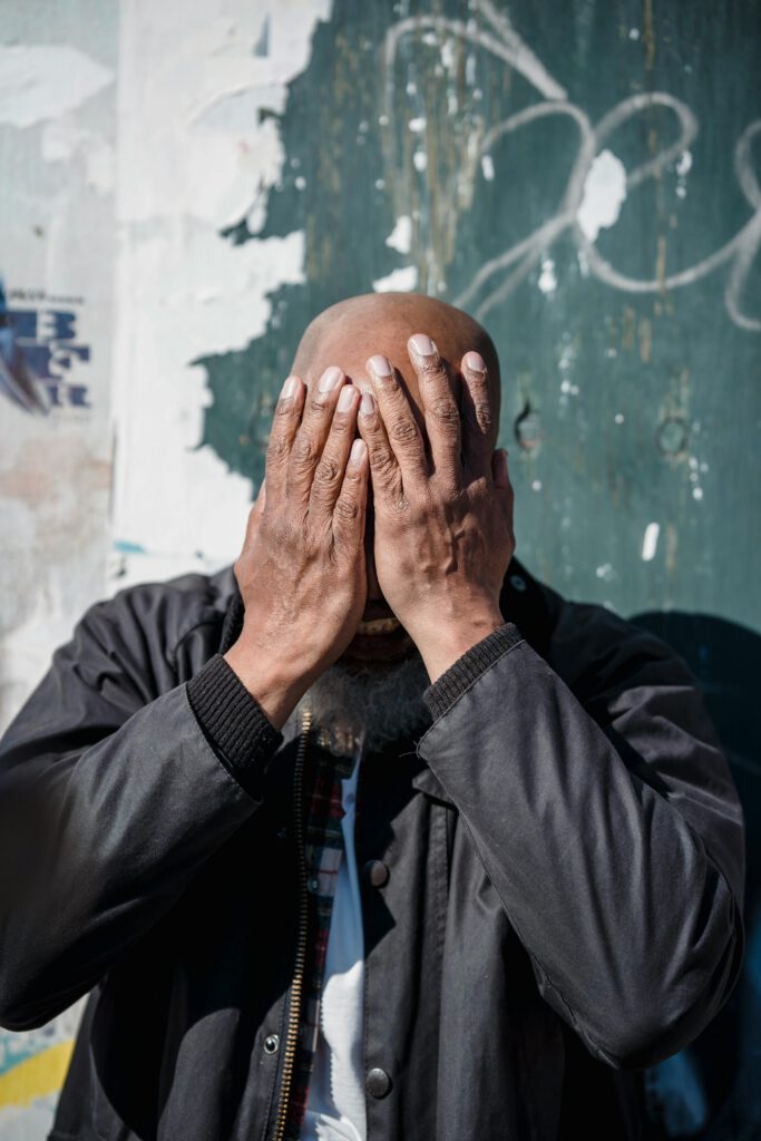 A bald man expresses frustration outdoors against a worn wall, covering his face with his hands