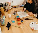 Artist painting a mandala design using various pigments and brushes indoors.